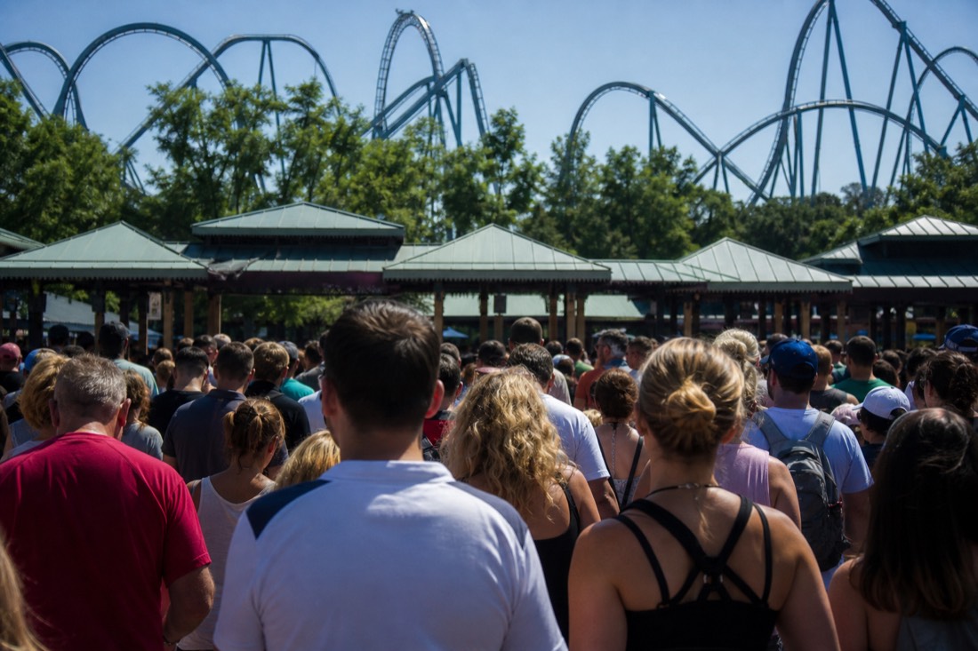Late-morning crowd bunched at a theme-park entrance gate under a clear sky, the cost of a slow start